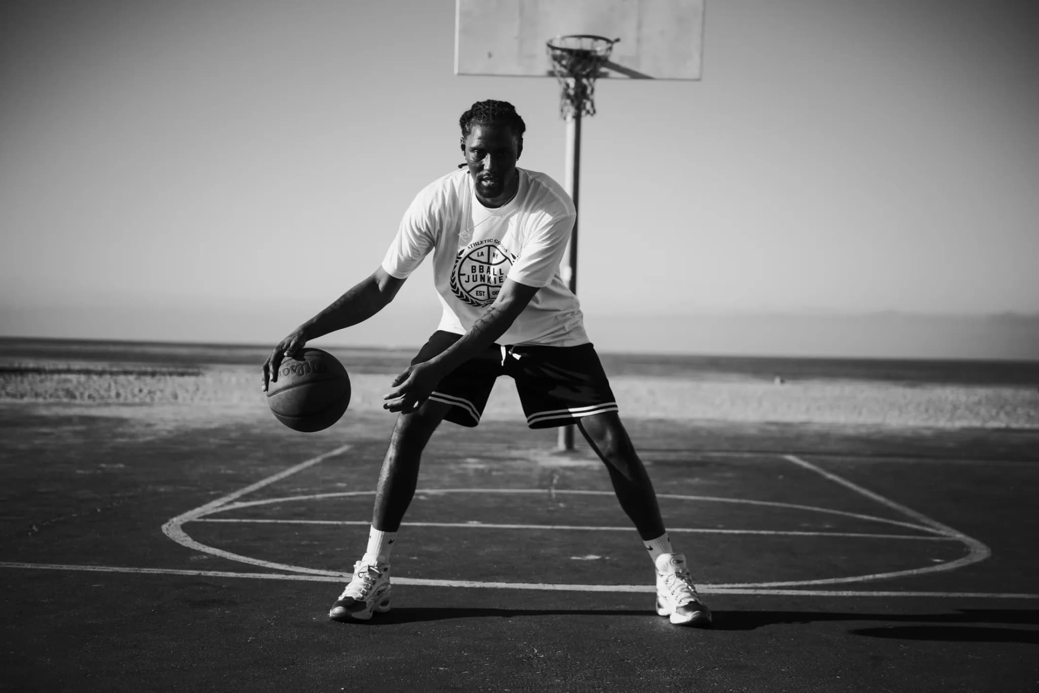 A player squared up with the ball at his hip under a beachside hoop, in a white BBJ crest tee and black shorts.