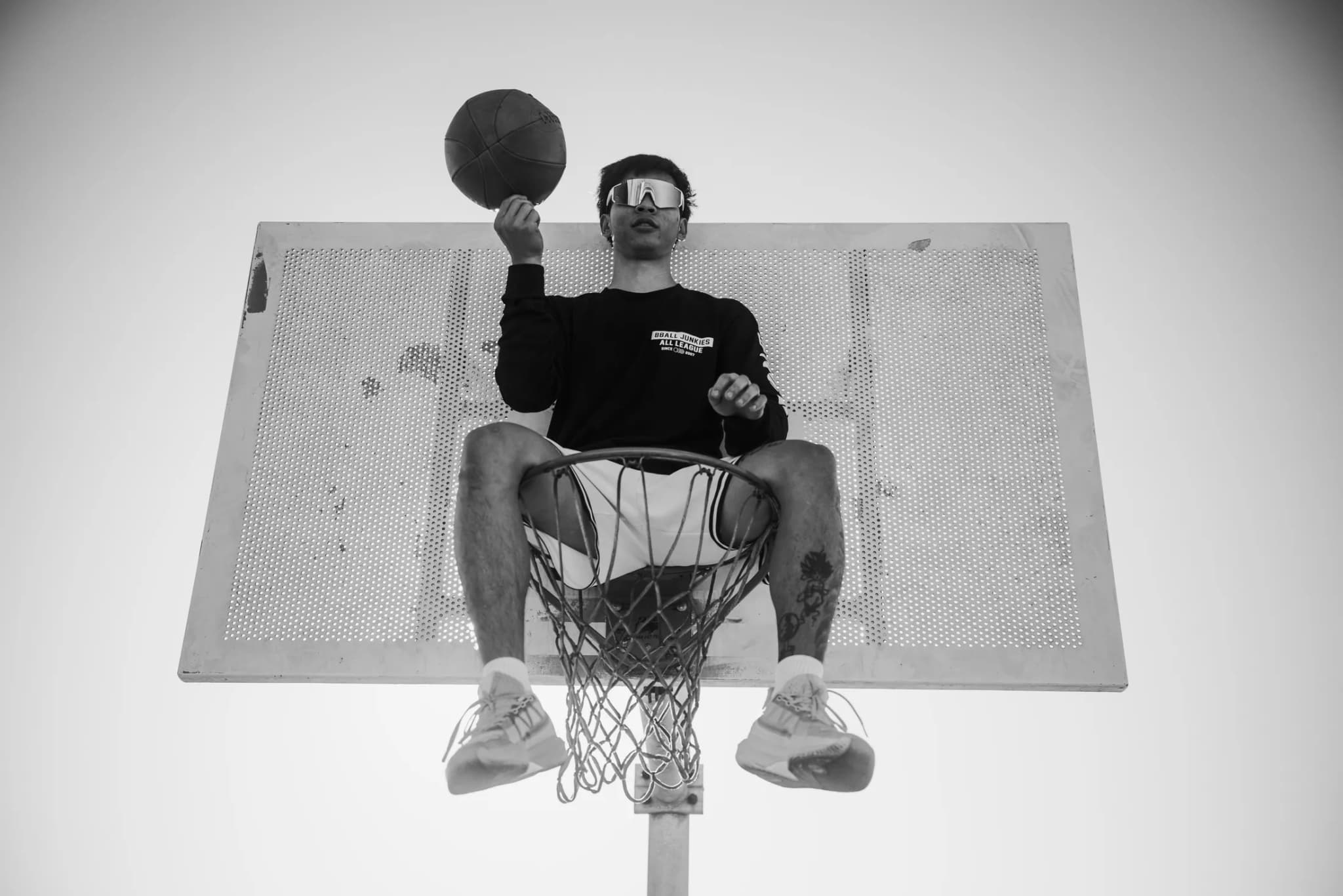 A player sitting in the hoop itself, legs through the net, spinning a ball on one finger — shot from directly below the backboard.