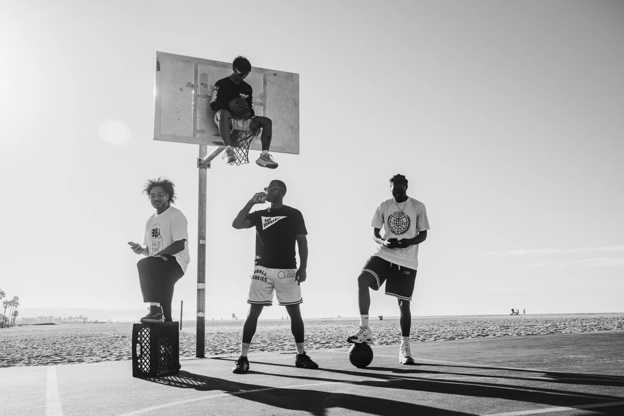 Three players on a beachside court, one perched up on the backboard, the two below with a ball and a milk crate.