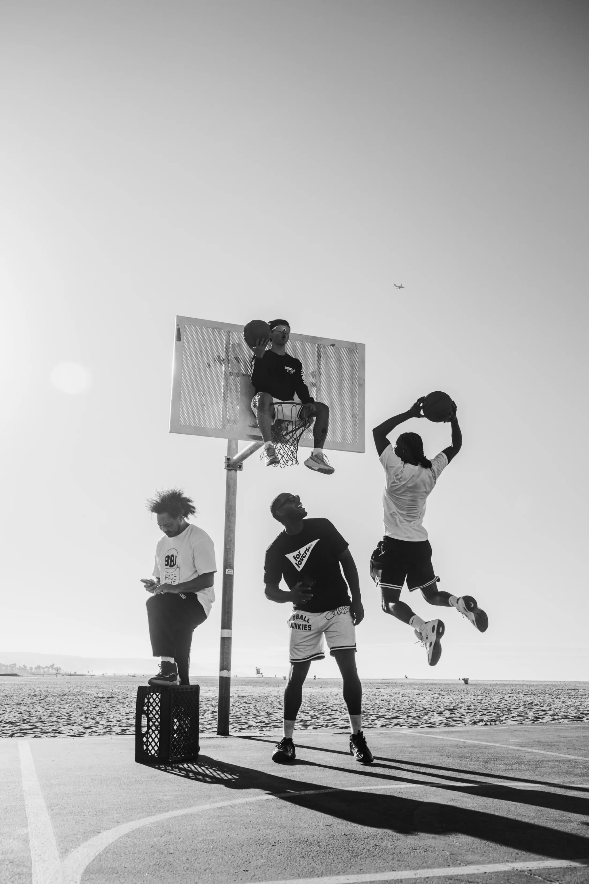 Three players under a beachside hoop, one perched on the backboard, one rising for a dunk against the sky.