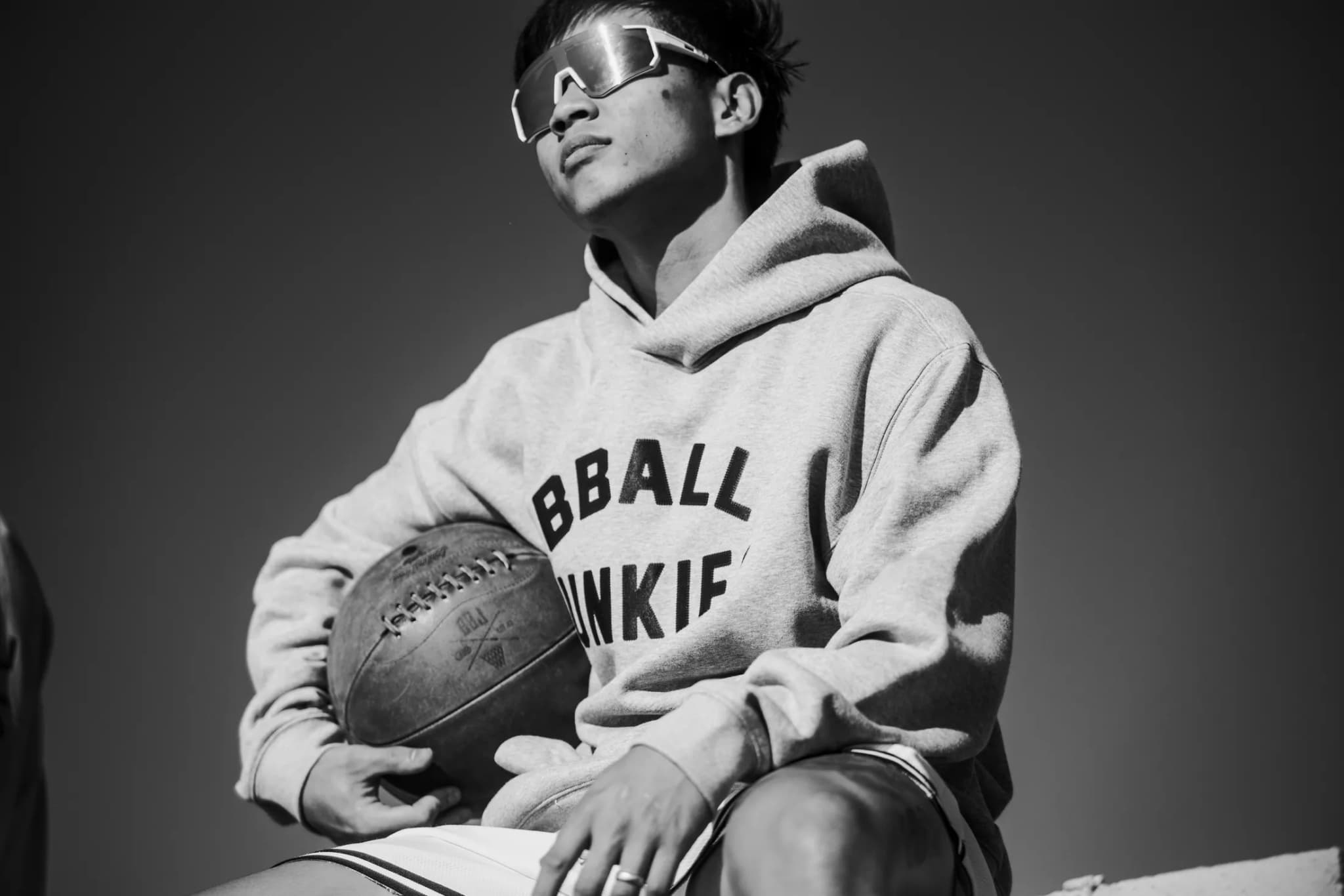 A player in the heather-gray BBALL JUNKIES hoodie and white shield sunglasses, sitting with a leather ball in one arm, shot from below against a clear sky