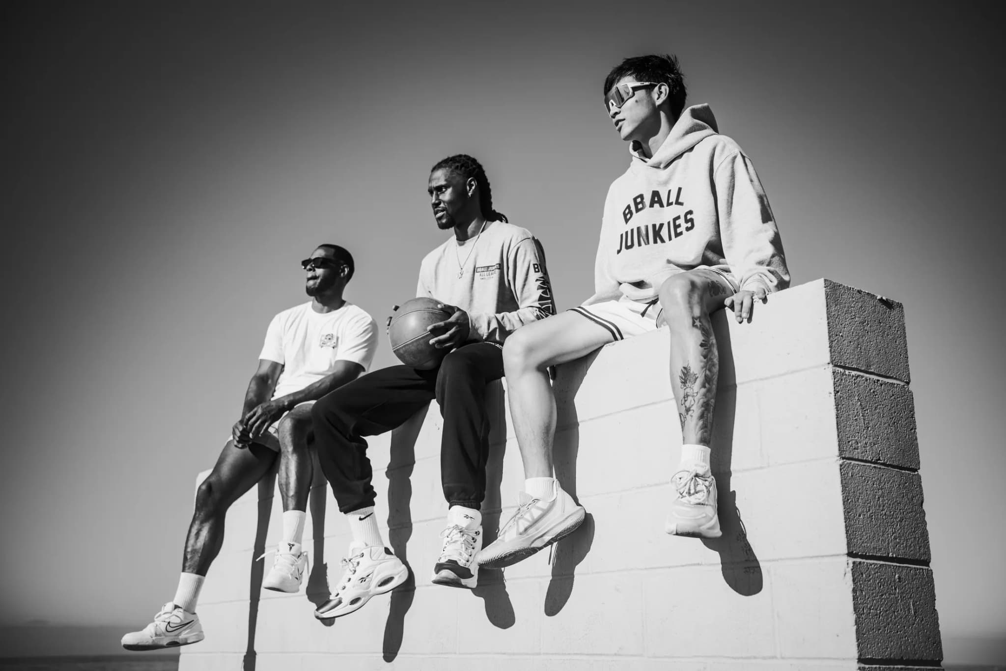 Three players sitting on a block wall against the sky, in BBJ tees and the gray BBALL JUNKIES hoodie, one holding a ball.
