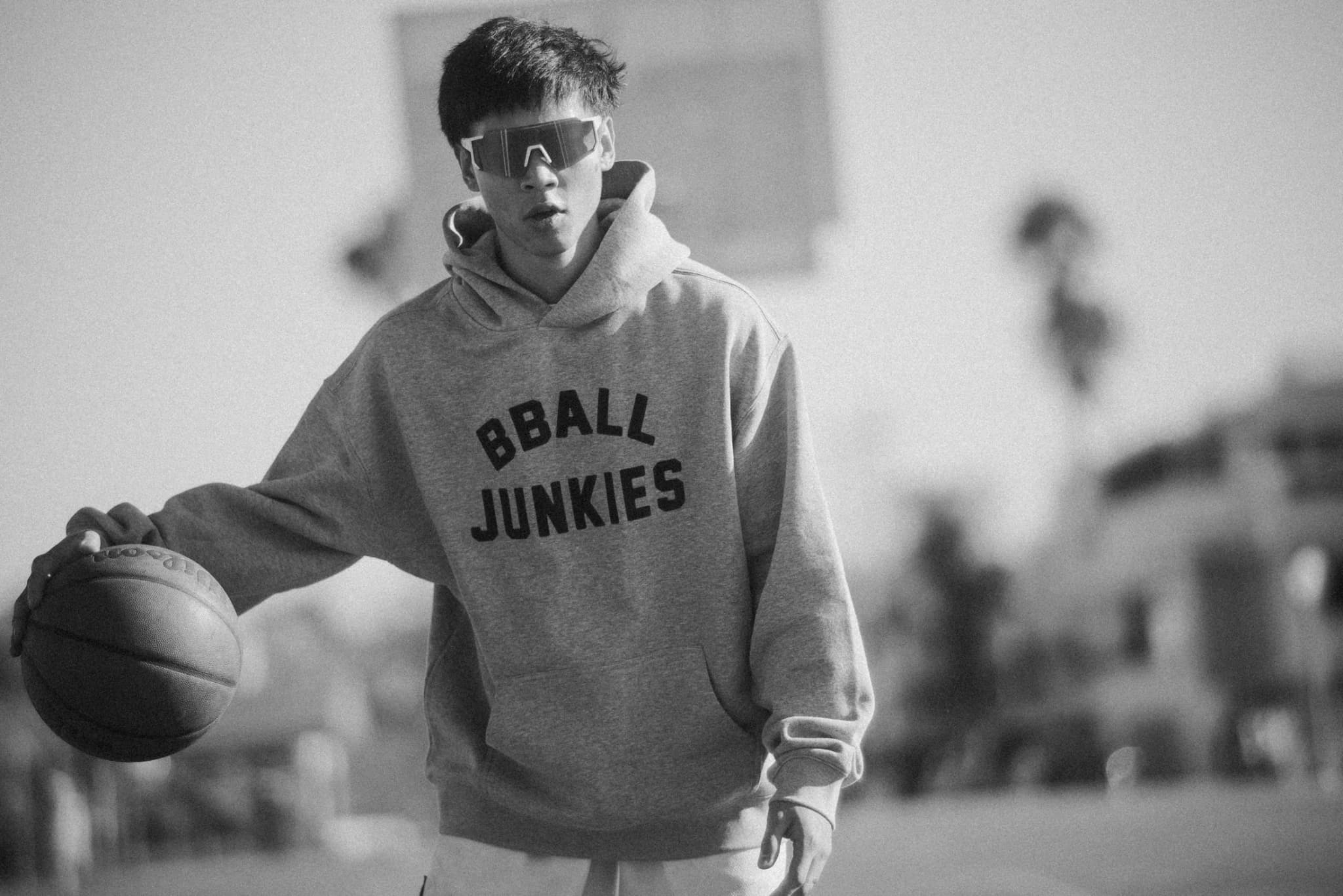 A player in mirrored shades dribbling toward the camera in the gray BBALL JUNKIES hoodie, palm trees soft behind him.