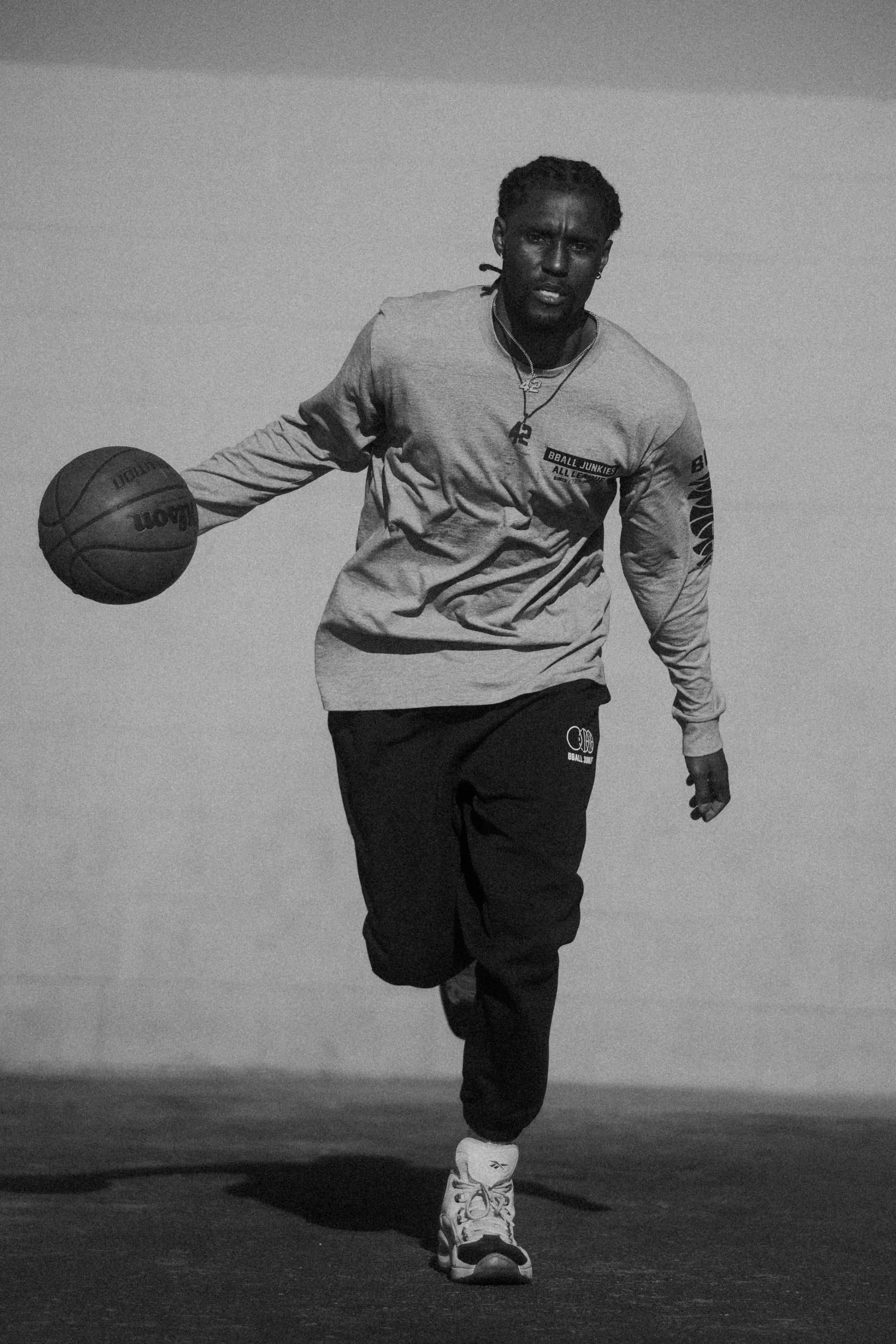 A player dribbling toward the camera along a white block wall, in a gray long sleeve and black pants.