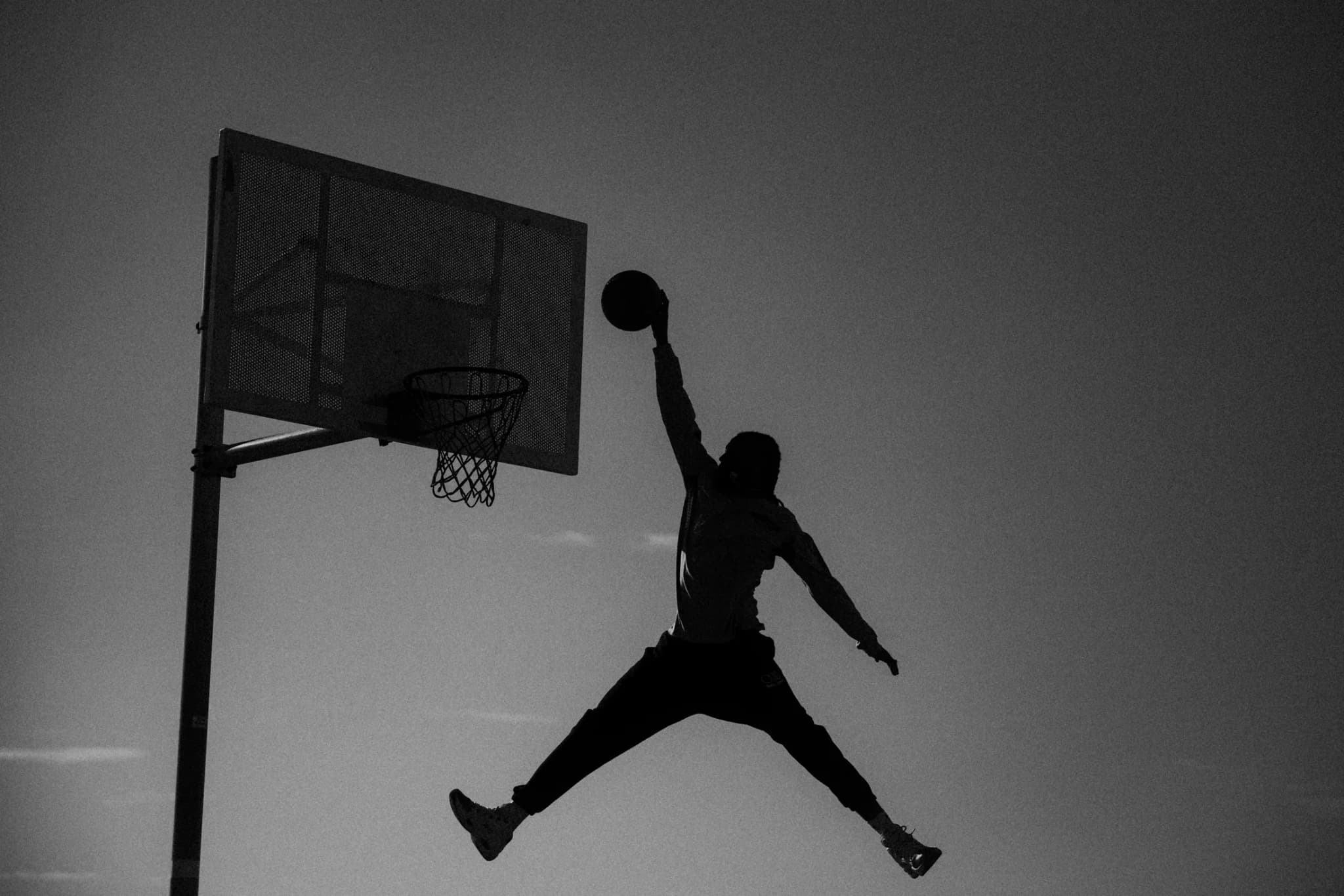A silhouette of a player rising for a one-handed dunk on a beachside hoop, against a flat sky.