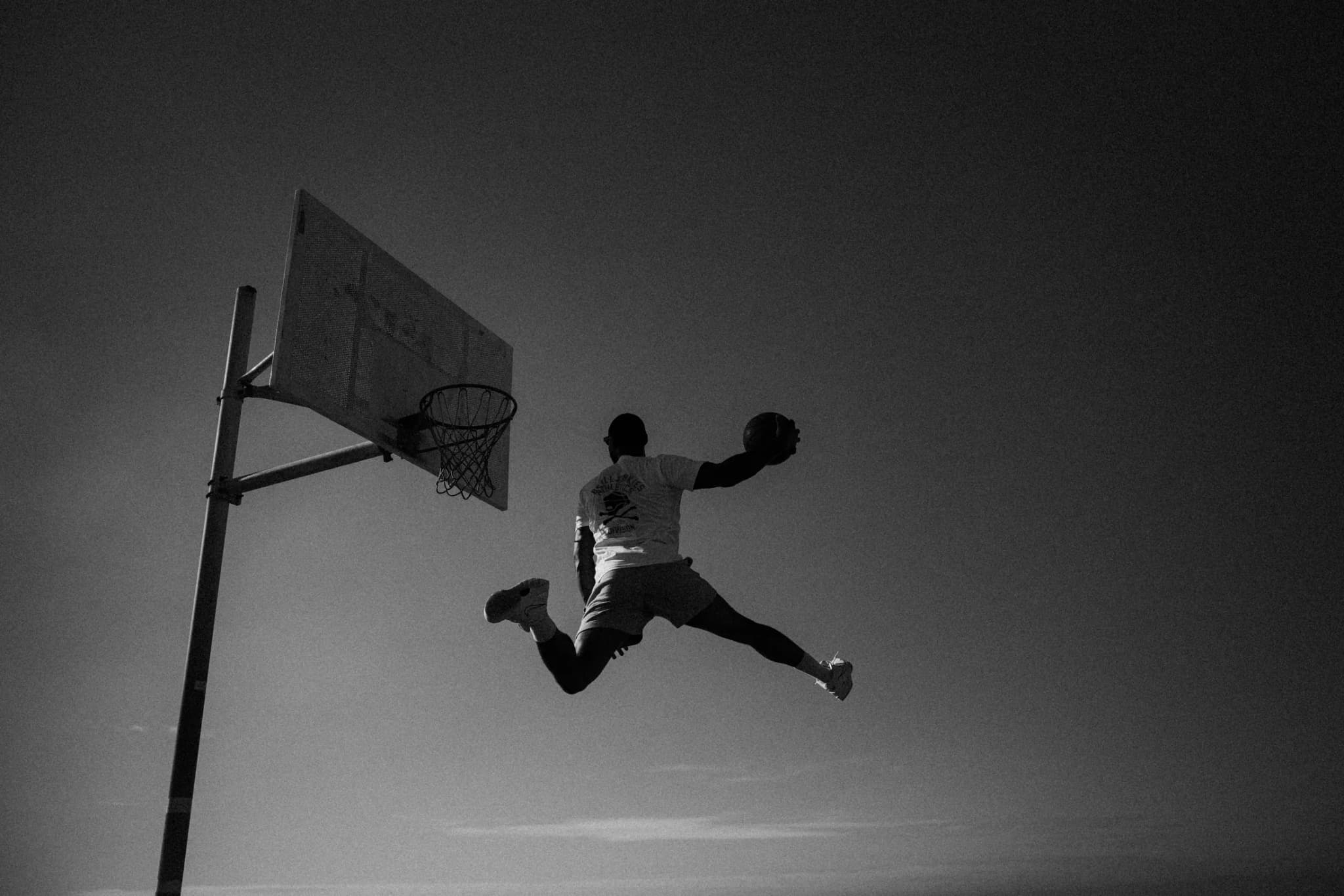 A player in mid-air with the ball in both hands, coming off a jump under a beachside hoop against the sky.