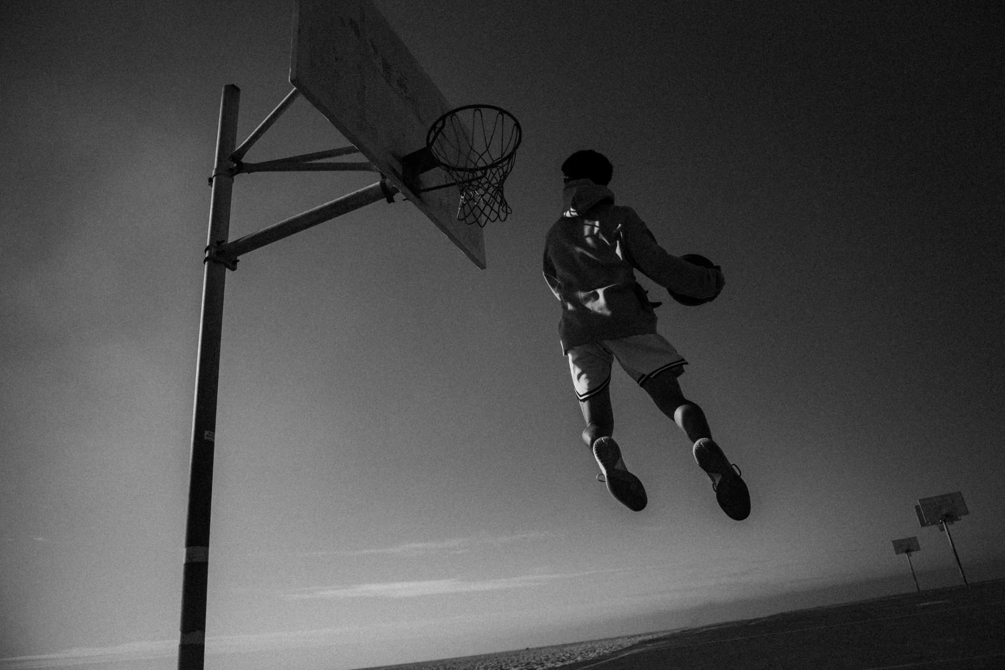 A player rising toward the rim with the ball, shot from below against a clear sky, a second hoop on the horizon.