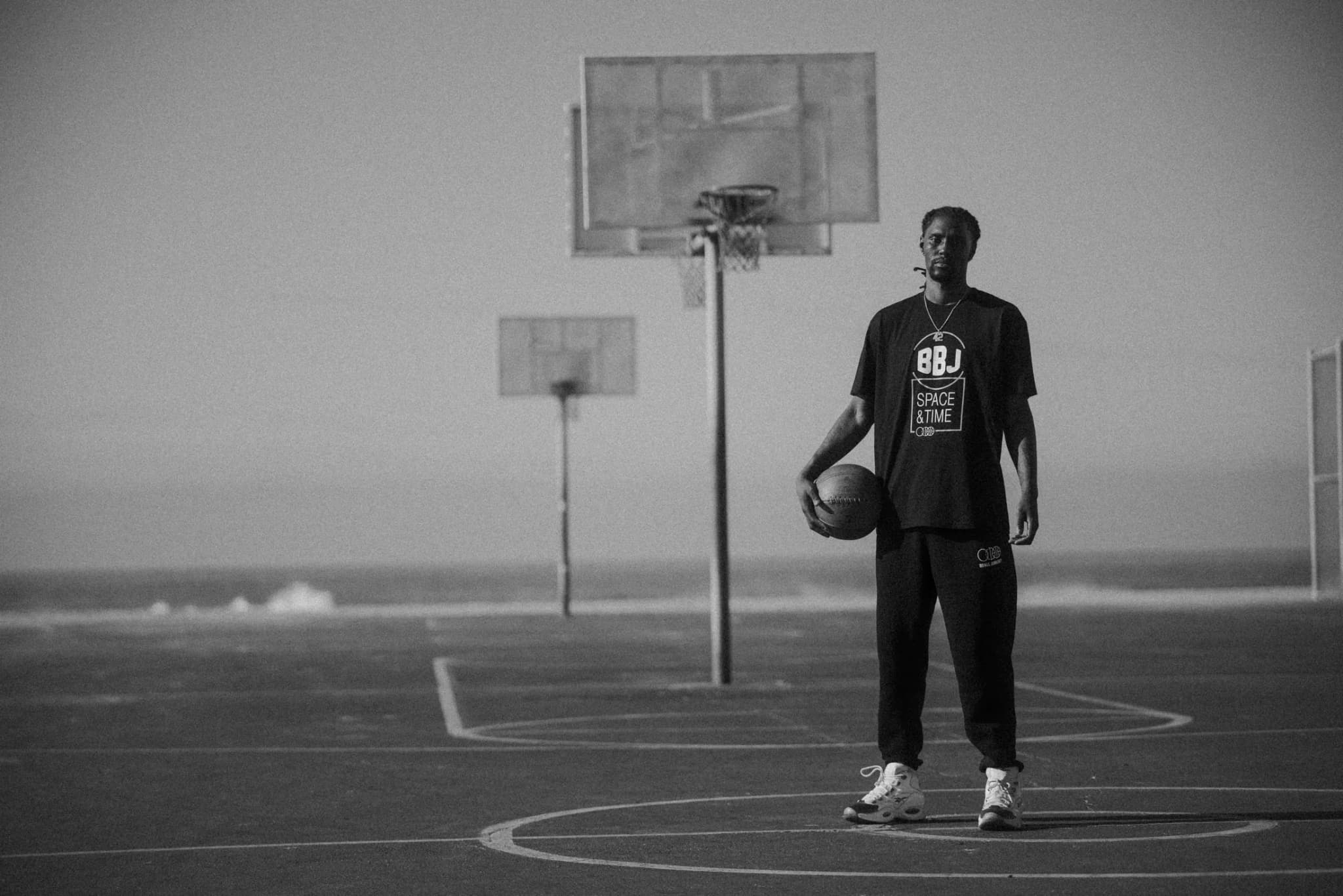 A player standing at the top of the key on a beach court with a ball on his hip, in the black BBJ Space & Time tee and sweats, three backboards and the ocean behind him