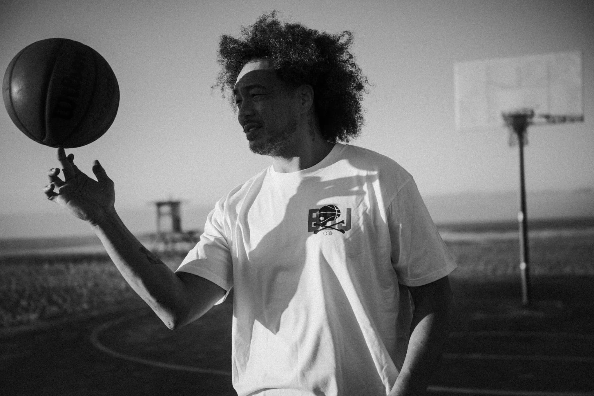 A player spinning a ball on one finger on a beach court, looking off to the side, in a white tee with the BBJ crest on the chest