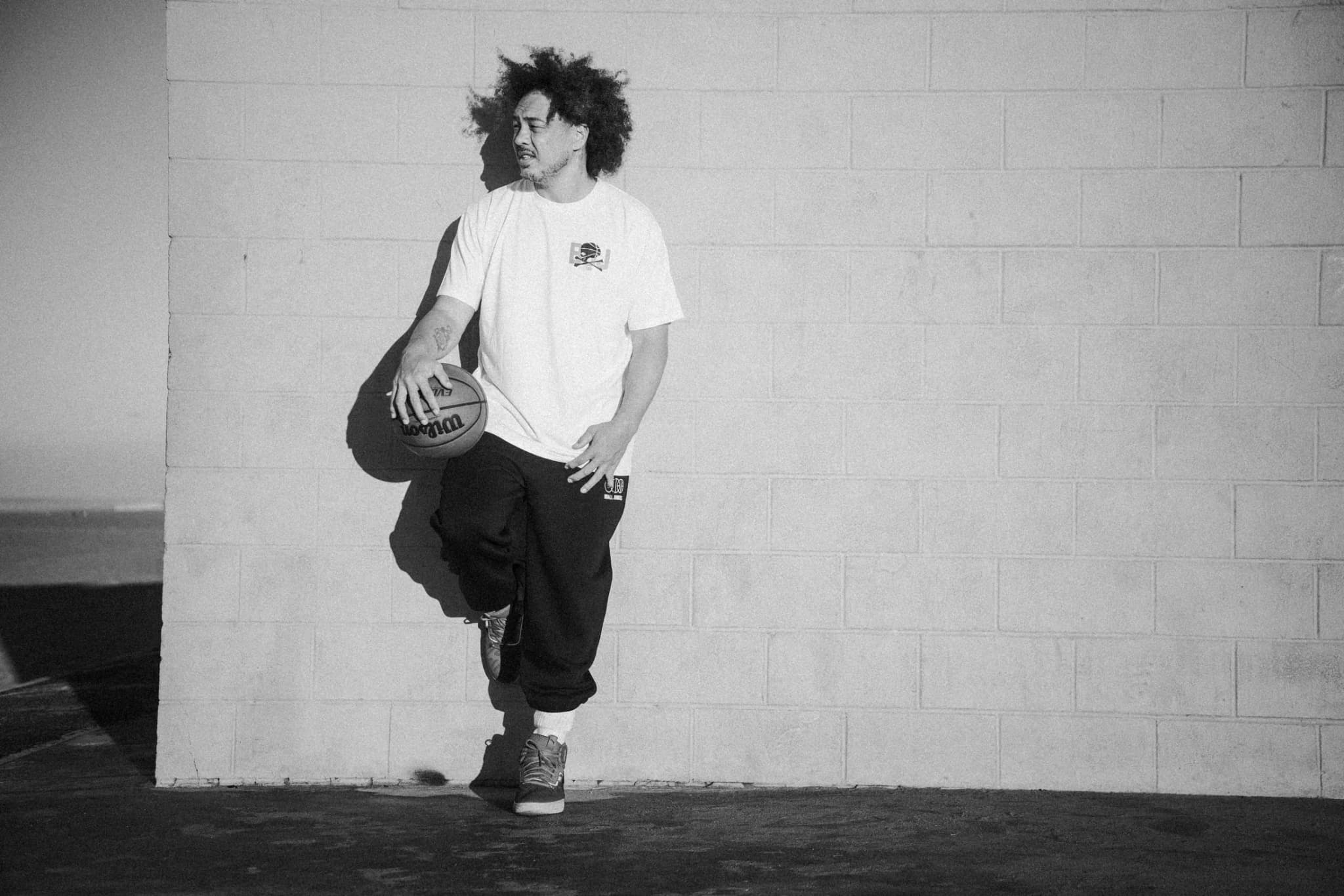 A player leaning one foot up against a white block wall with a ball under his arm, in a white BBJ tee and dark pants.
