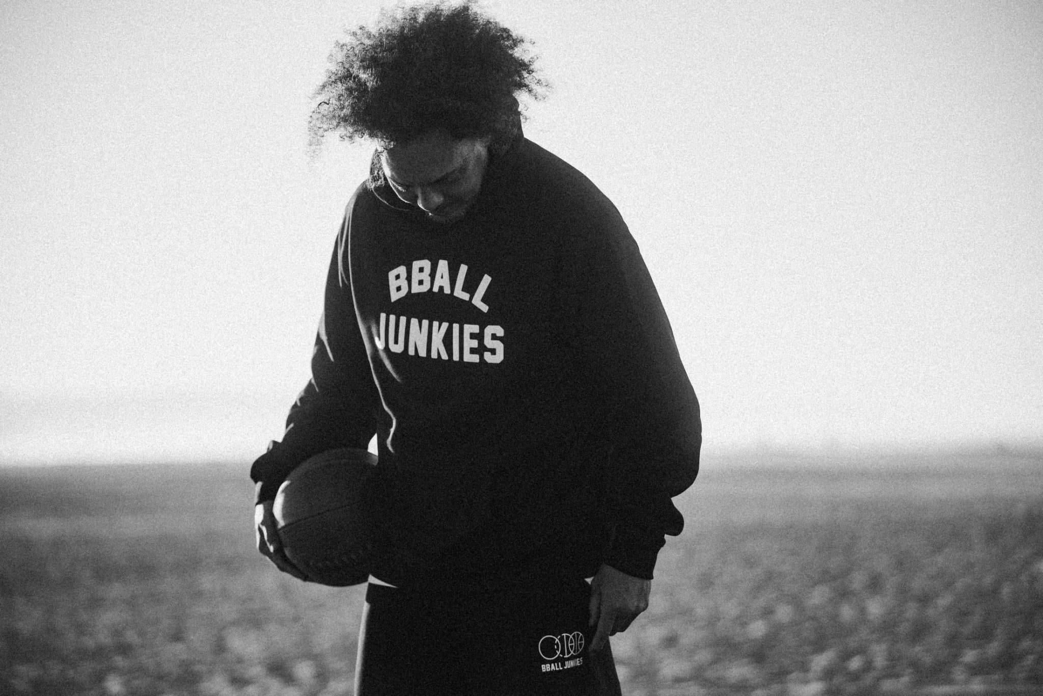 A player looking down at the ball in his hands, in a black BBALL JUNKIES crewneck, the sand and the sea soft behind him.