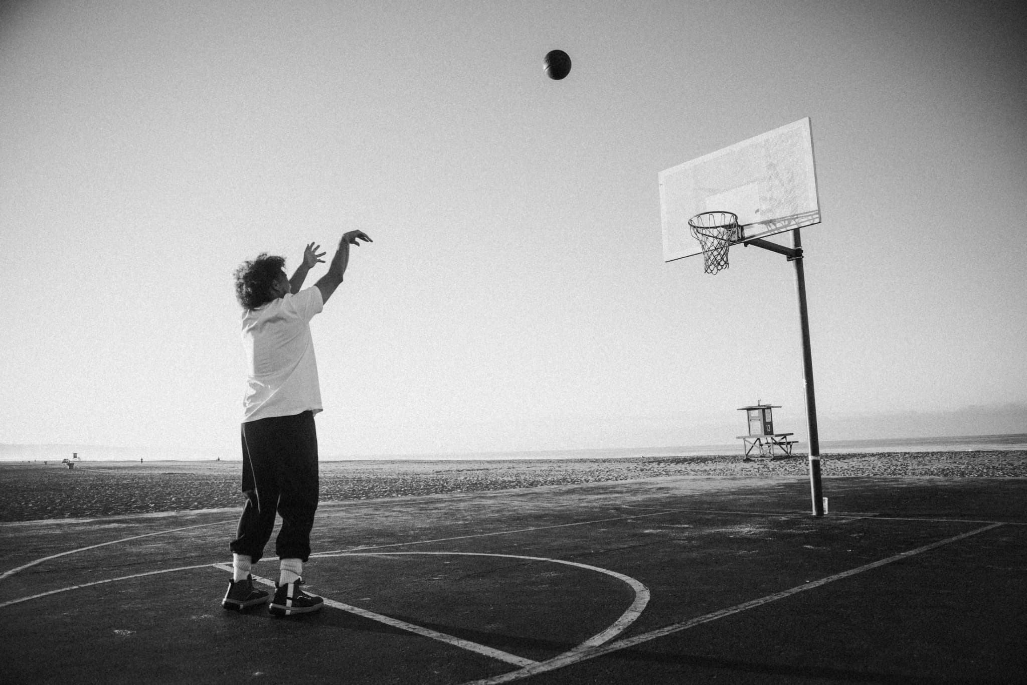 A lone player following through on a jump shot toward a beachside hoop, the ball high in the air, a lifeguard tower on the sand beyond.