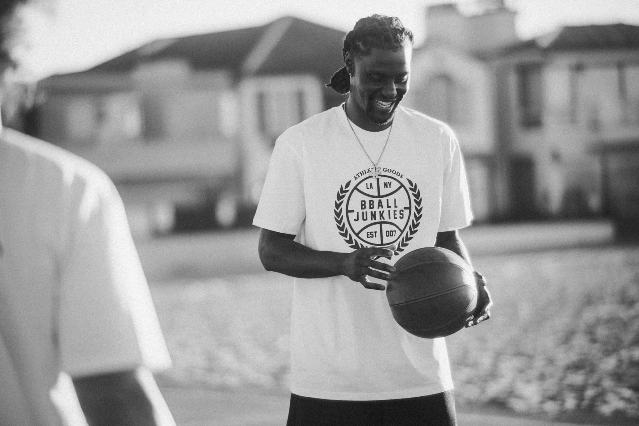 A player laughing down at the ball in his hands, in the white BBALL JUNKIES Athletic Goods laurel tee, LA NY and Est. 007 in the crest, houses soft behind him