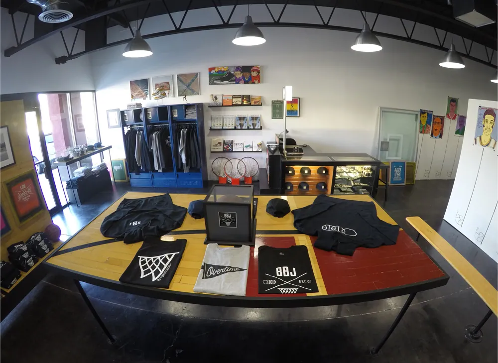 The Locker Room retail floor: tees laid out on a display table built from court parquet, lockers racked with garments, hoops and artwork along the walls.