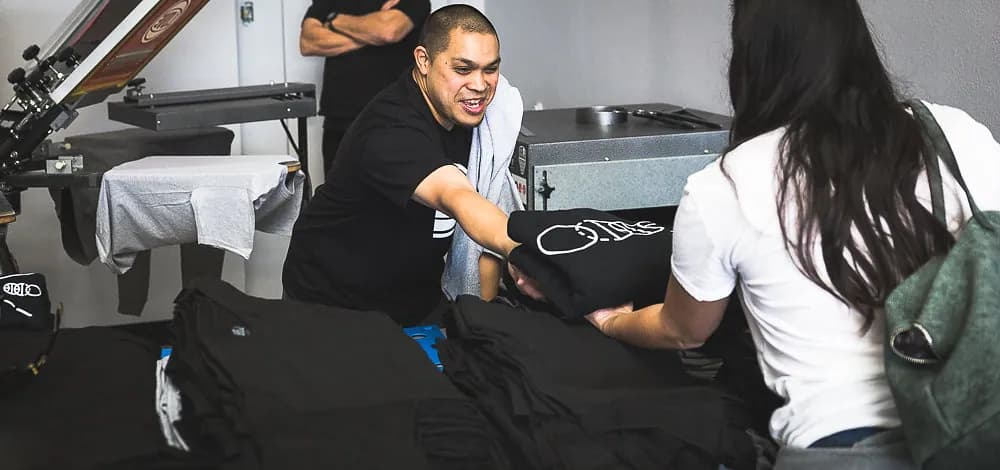 A folded, freshly printed tee handed across a table of finished shirts at the BBJ Locker Room, the carousel press and heat press behind.