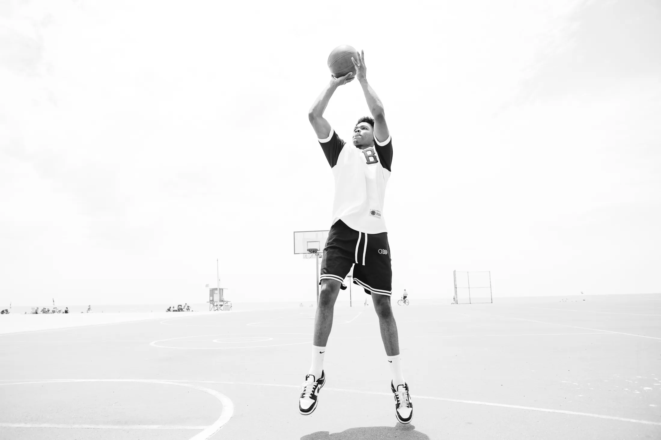 A player rising into a jump shot on a seaside court, in a white BBJ shirt with a chenille B on the chest and black mesh shorts.