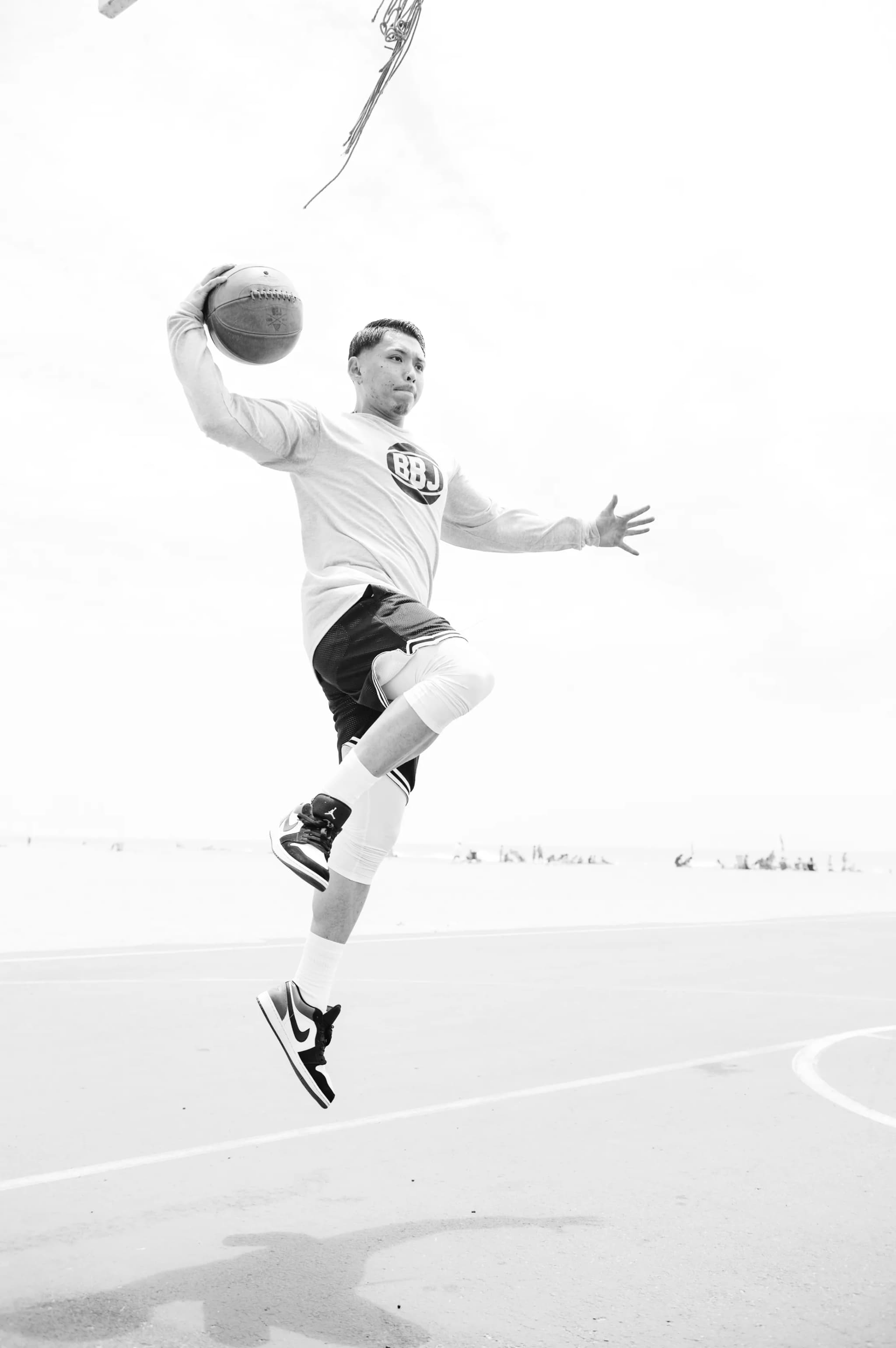 A player in the air with the ball cocked behind his head, in a long-sleeve BBJ tee and mesh shorts, the rim above him.