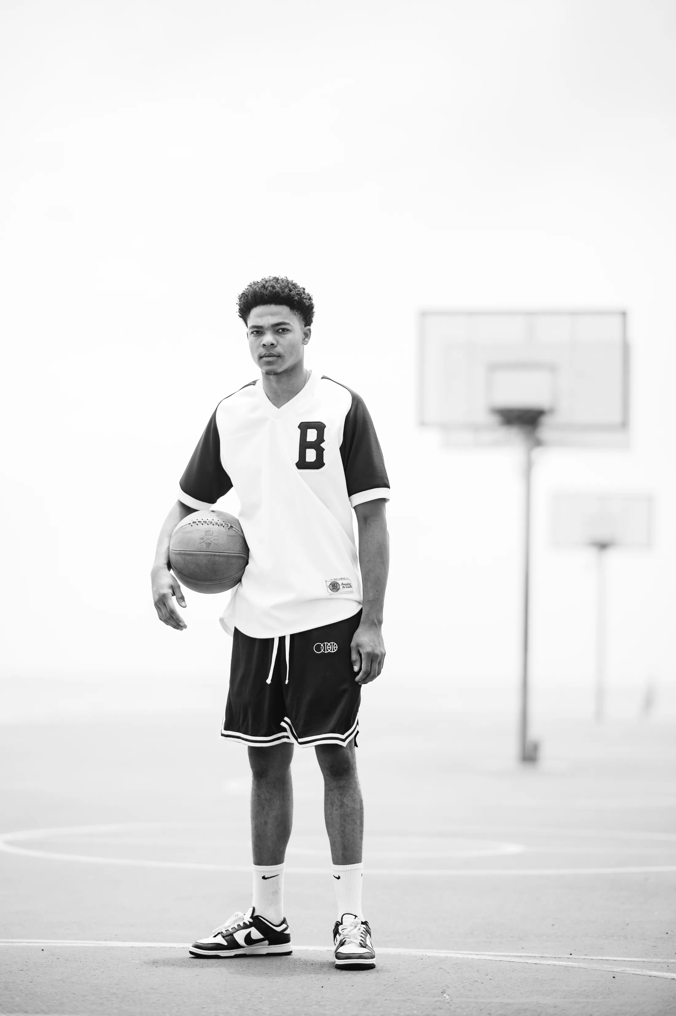 A player standing on an outdoor court holding a ball, in a BBJ B-logo jersey, backboards behind him.