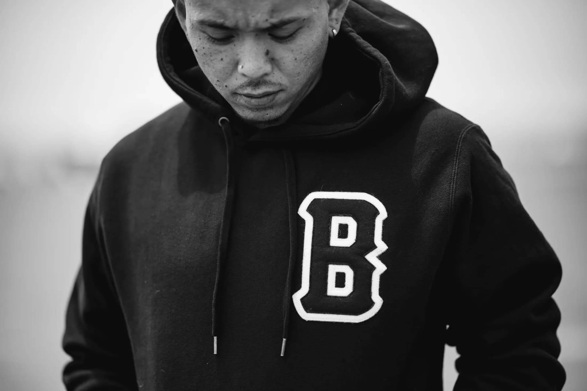 The chenille B hoodie worn head-down against a flat sky, both hands at the drawstrings.