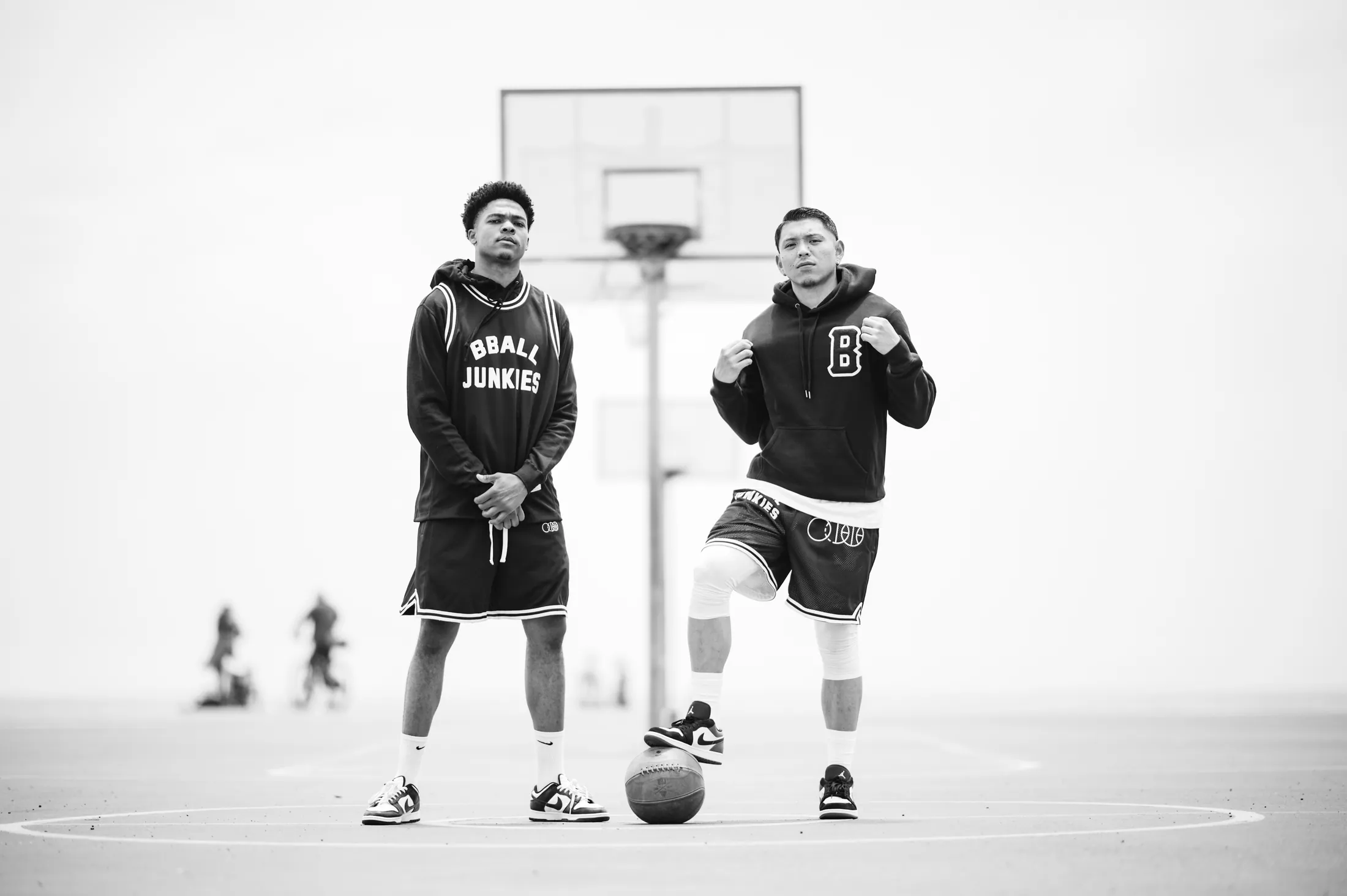 Two players under a backboard on an outdoor court, one in a BBALL JUNKIES warm-up jacket and mesh shorts, the other in a B-logo hoodie, a worn leather ball on the ground between them.