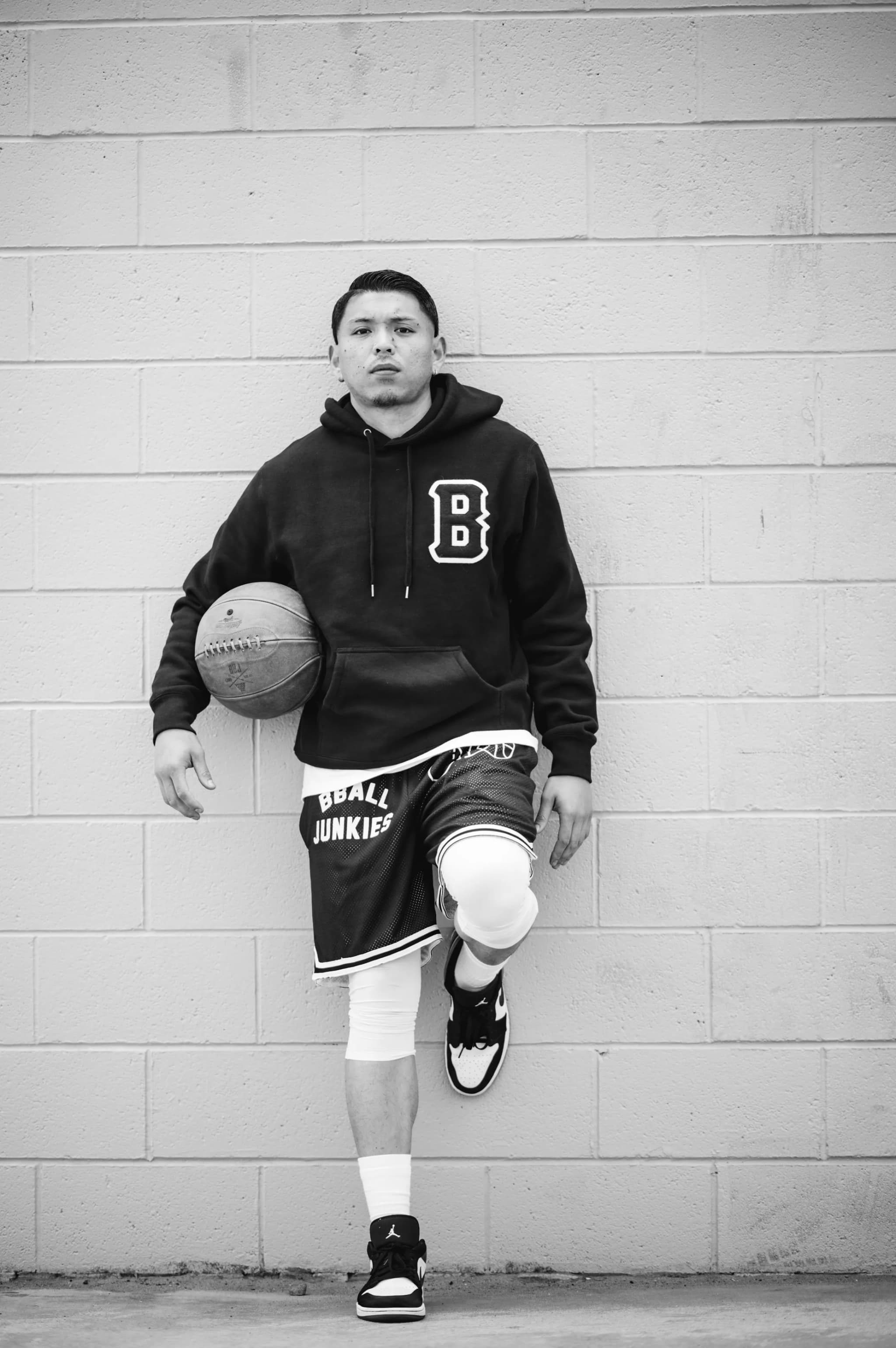 A player leaning against a cinder-block wall with a ball under his arm, in a black B-logo hoodie and BBALL JUNKIES shorts.