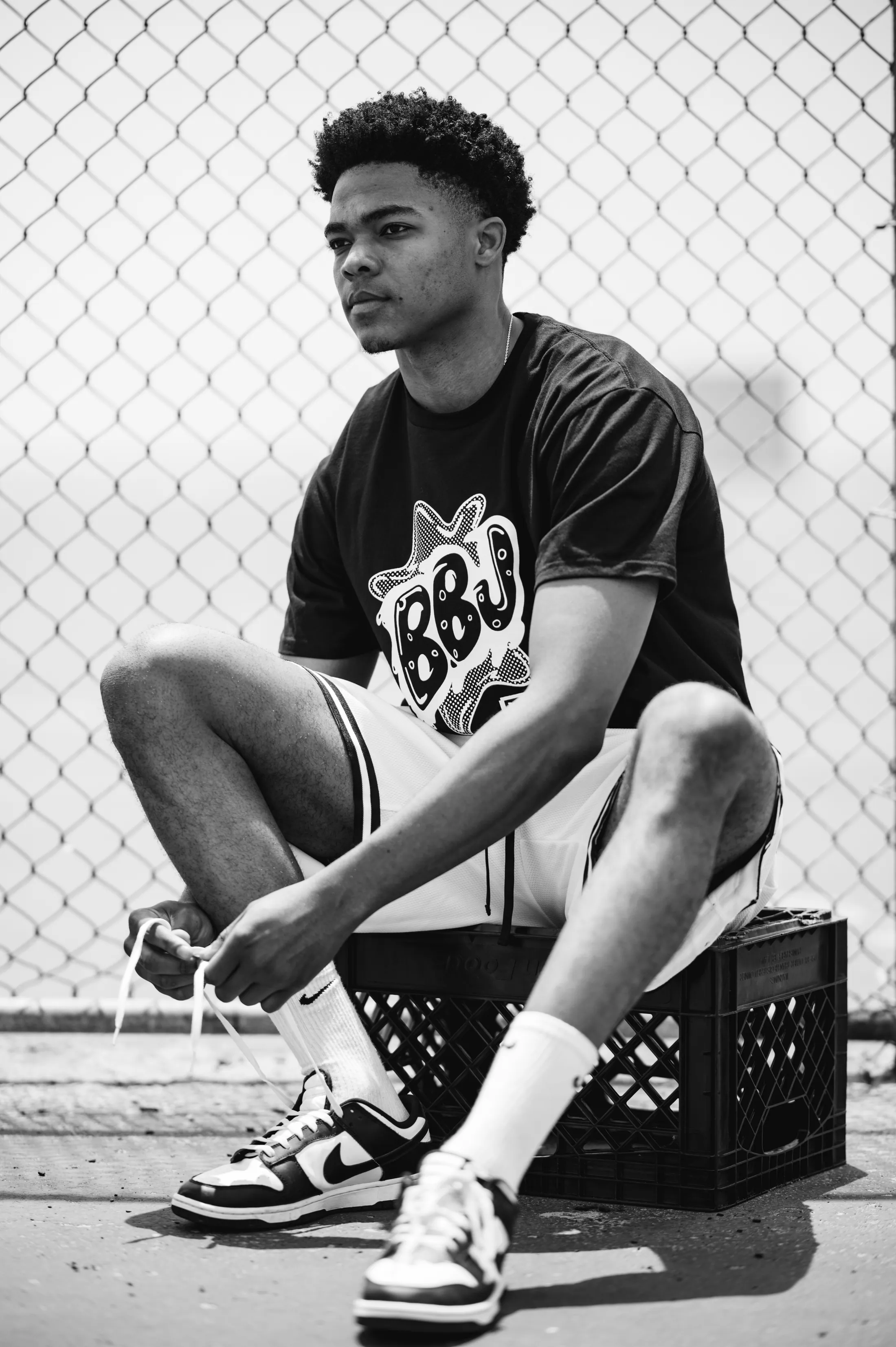 A player crouched on a milk crate against a chain-link fence, tying a shoe, in a BBJ tee.