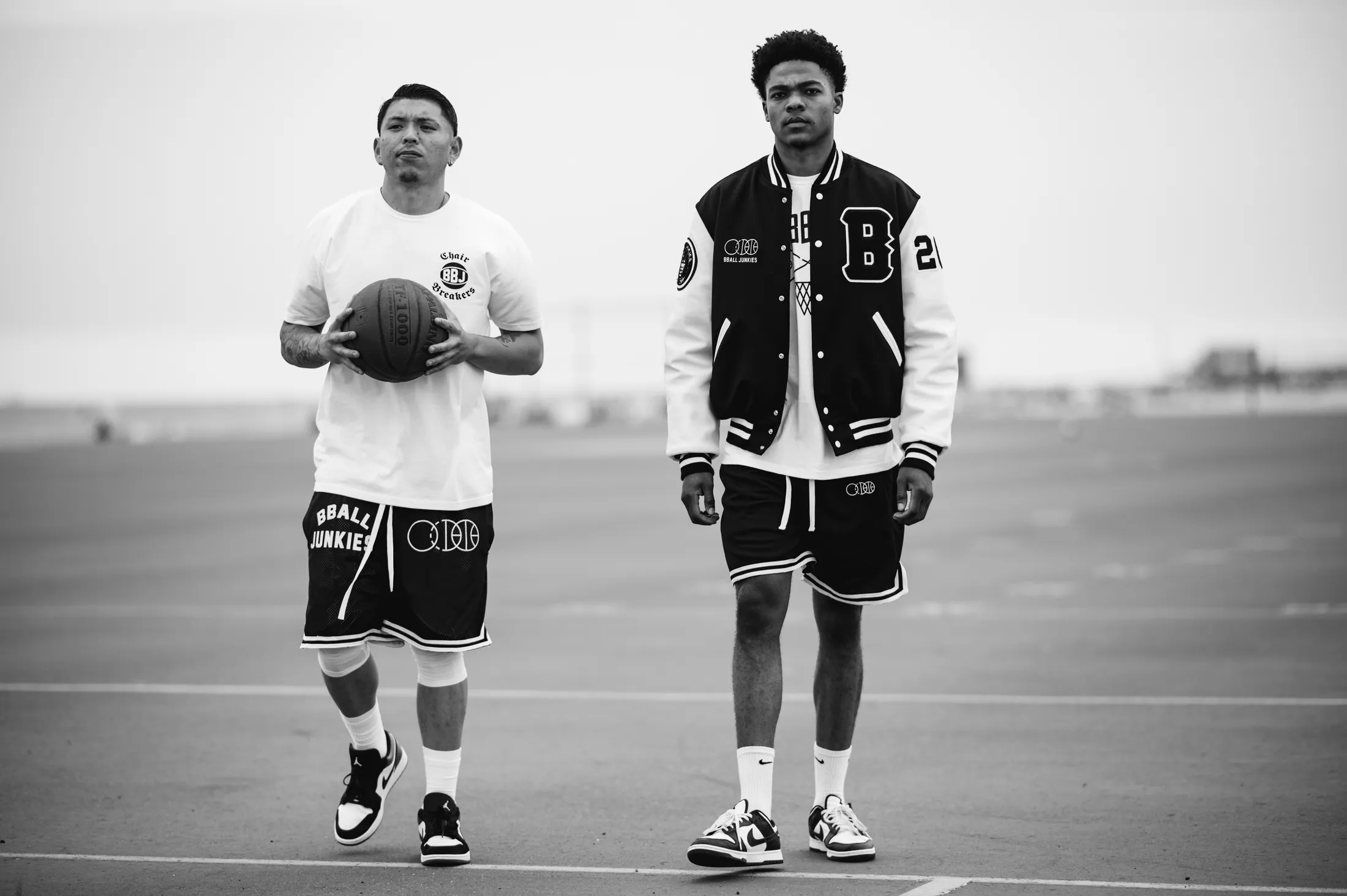 Two players walking across an empty court, one carrying a ball, one wearing the BBJ Authentic Varsity Jacket