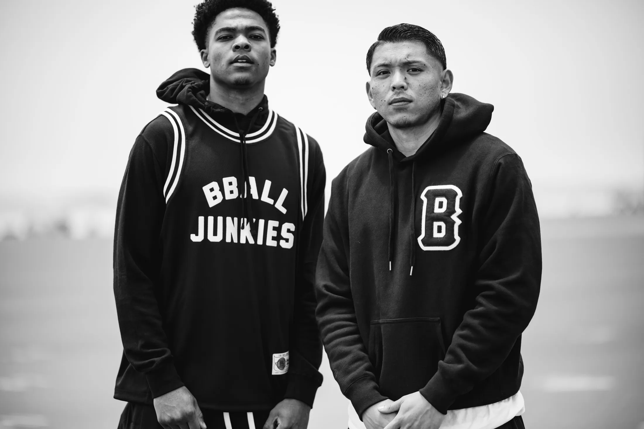 Two players standing together on an overcast beach, one in a BBALL JUNKIES warm-up jacket, the other in a hoodie carrying the chenille B.