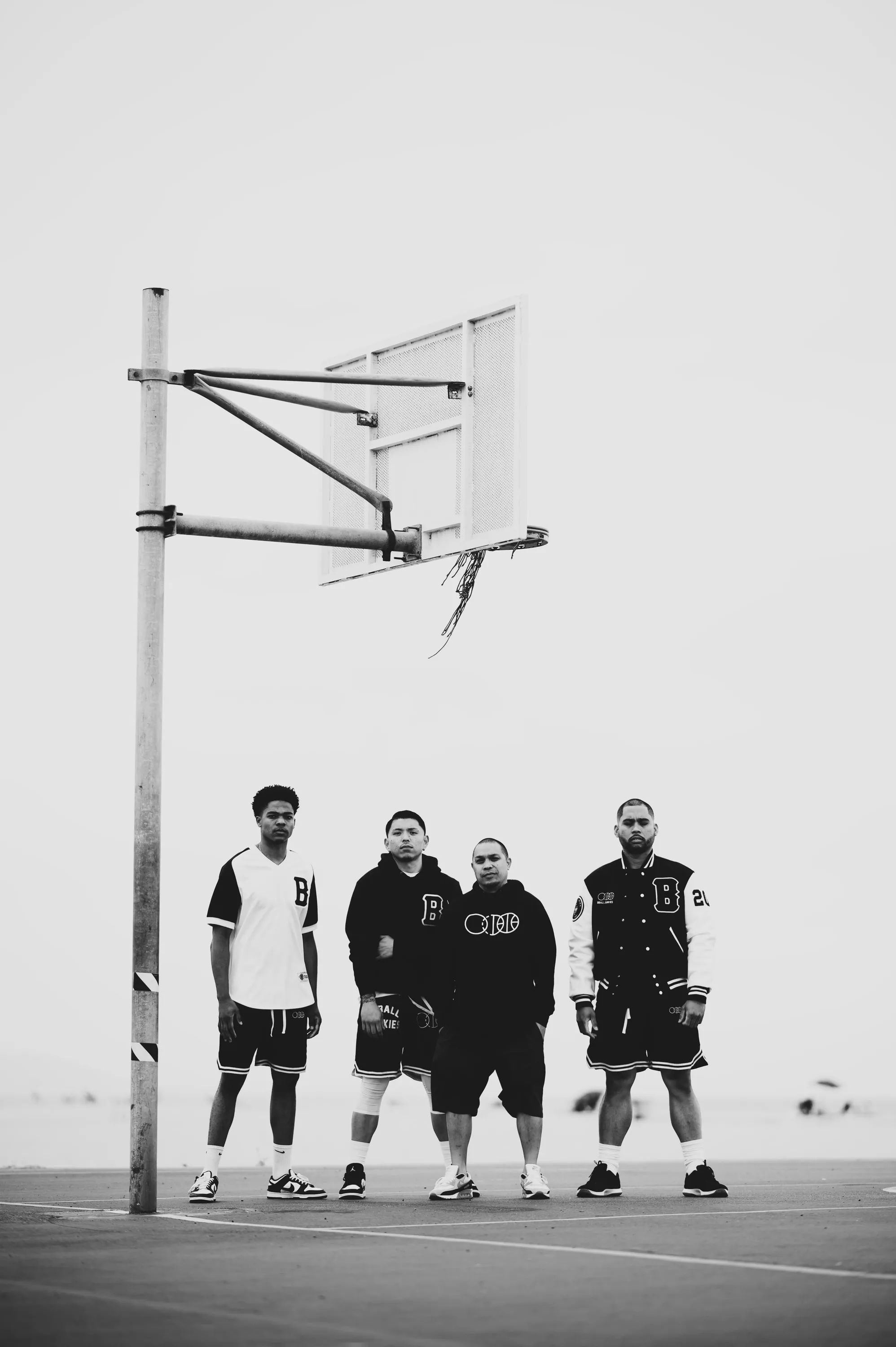 Four figures standing under a portable hoop against a blown-out white sky, in BBJ tees and crewnecks.