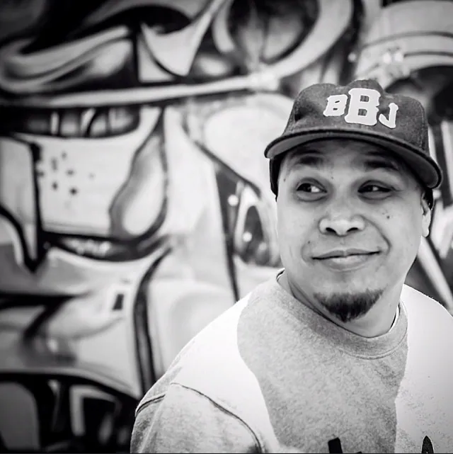 Rob Hernandez in a close black-and-white portrait in front of a graffiti wall, a BBJ cap worn low.