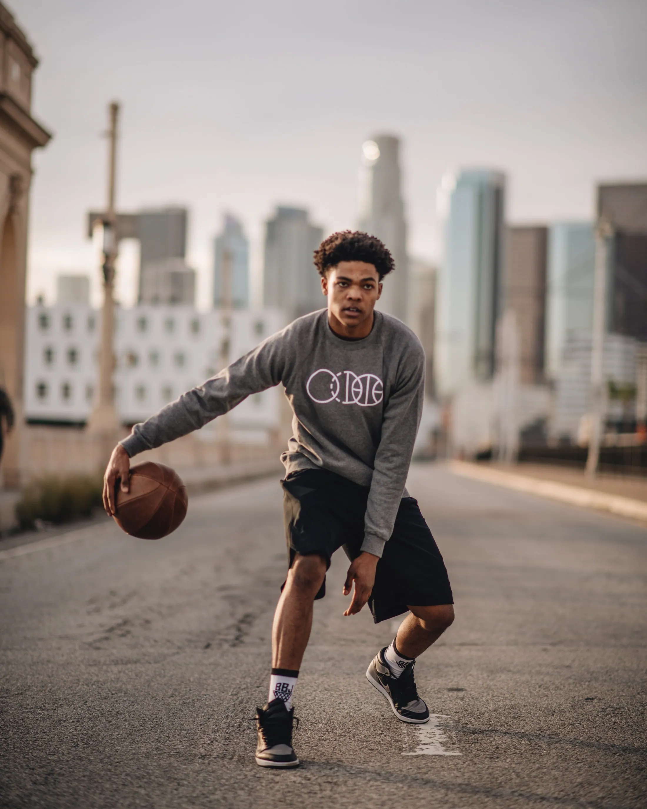 A player dribbling down an empty street with the downtown Los Angeles skyline behind him, in a gray BBJ crewneck.