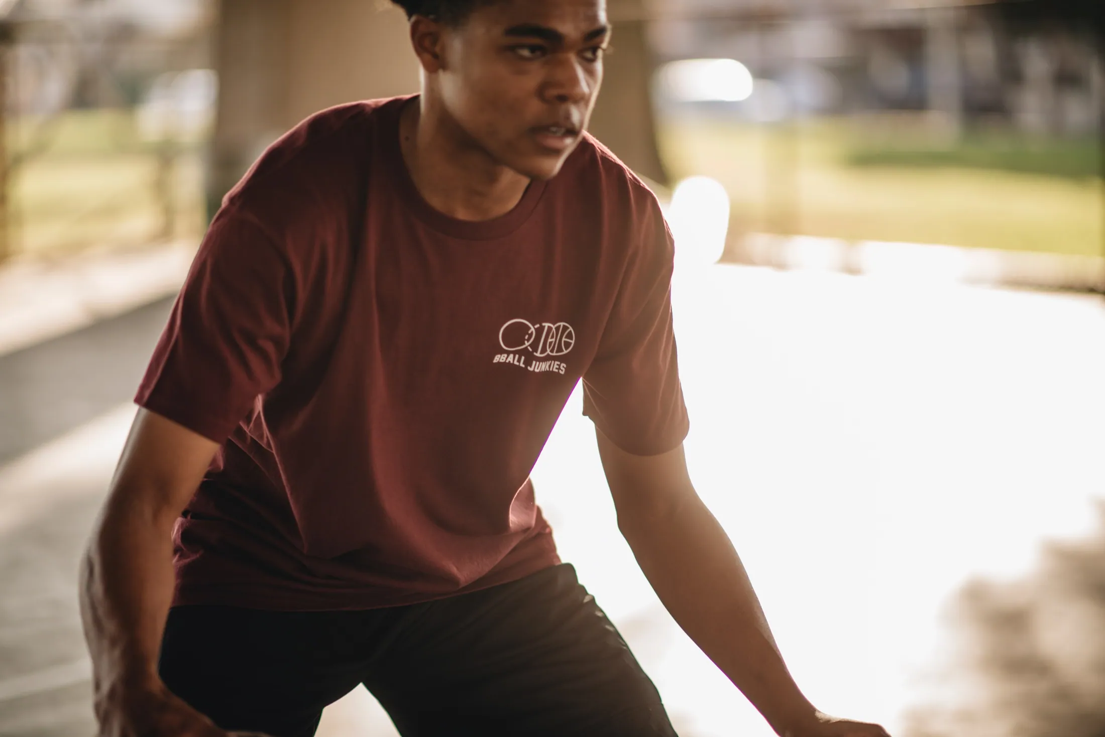 A player crouched into a defensive stance under a freeway underpass, in a burgundy tee carrying the BBJ three-ball mark.