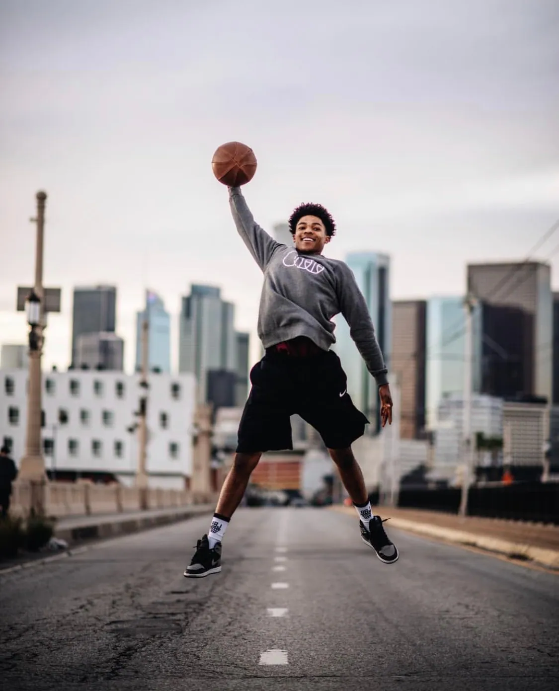 A player caught at the top of a jump on an empty street, ball held out at arm’s length, the Los Angeles skyline behind him.