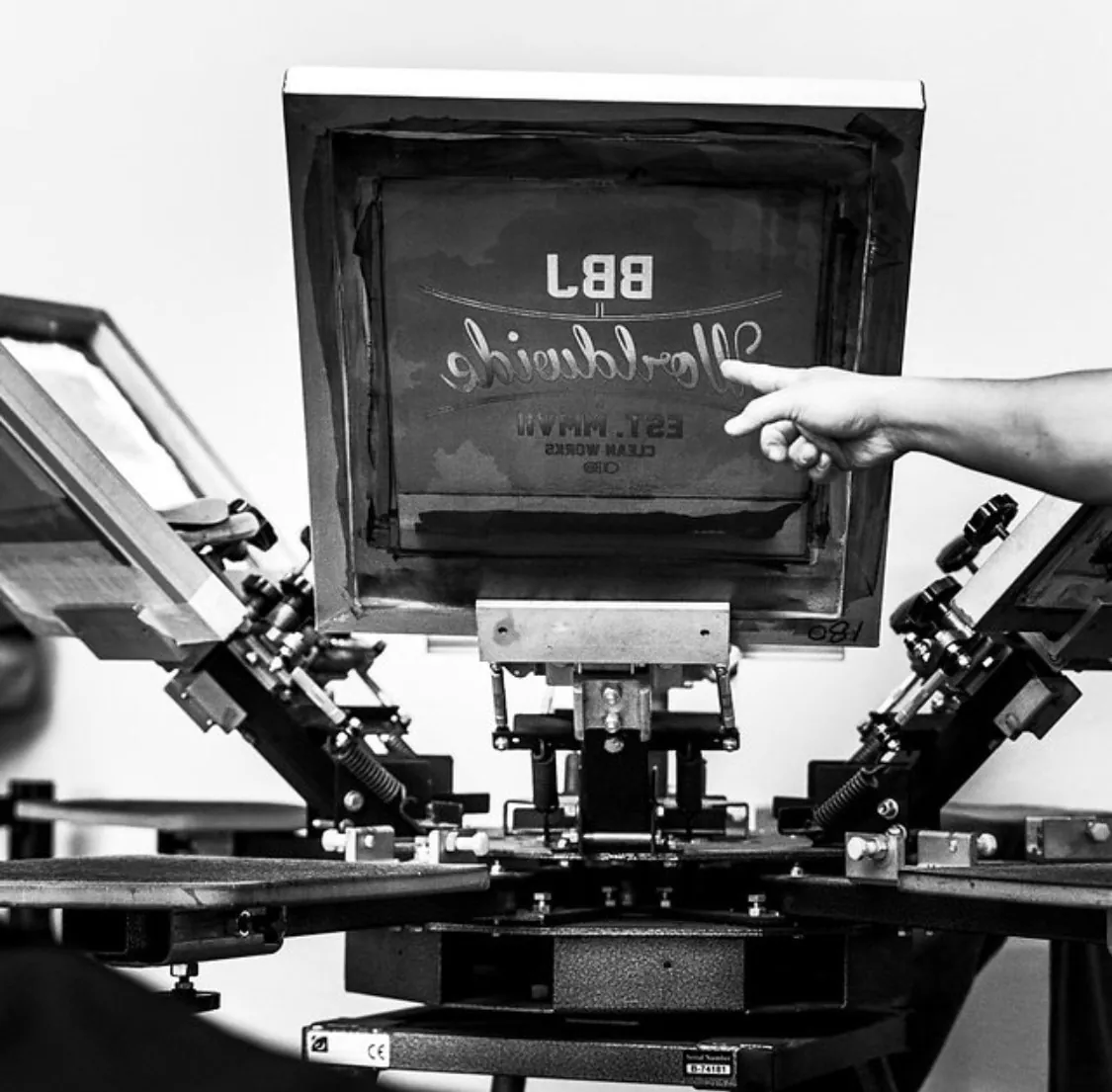 A hand resting on a burned screen racked over a carousel press, a customer's wordmark and est. date reading in reverse through the emulsion.