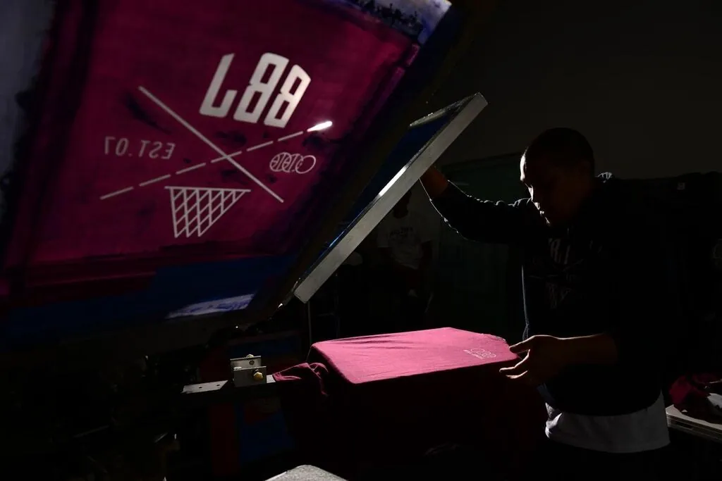 A printer lifting the screen off a freshly printed shirt on the press, the BBJ mark showing in reverse on the underside of the screen above.