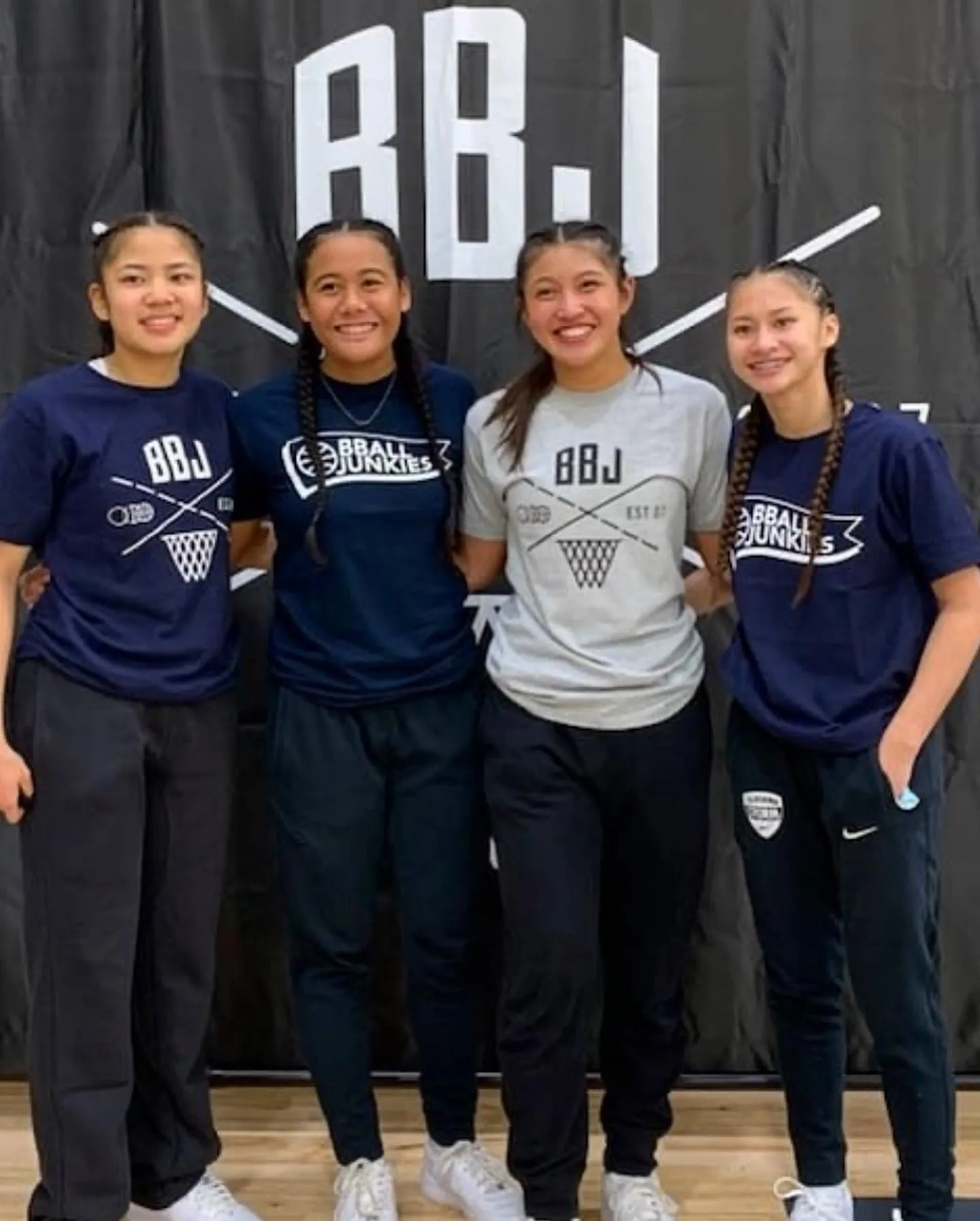 Four players in navy and gray BBJ tees lined up arm in arm on a gym floor in front of a black BBJ backdrop banner.