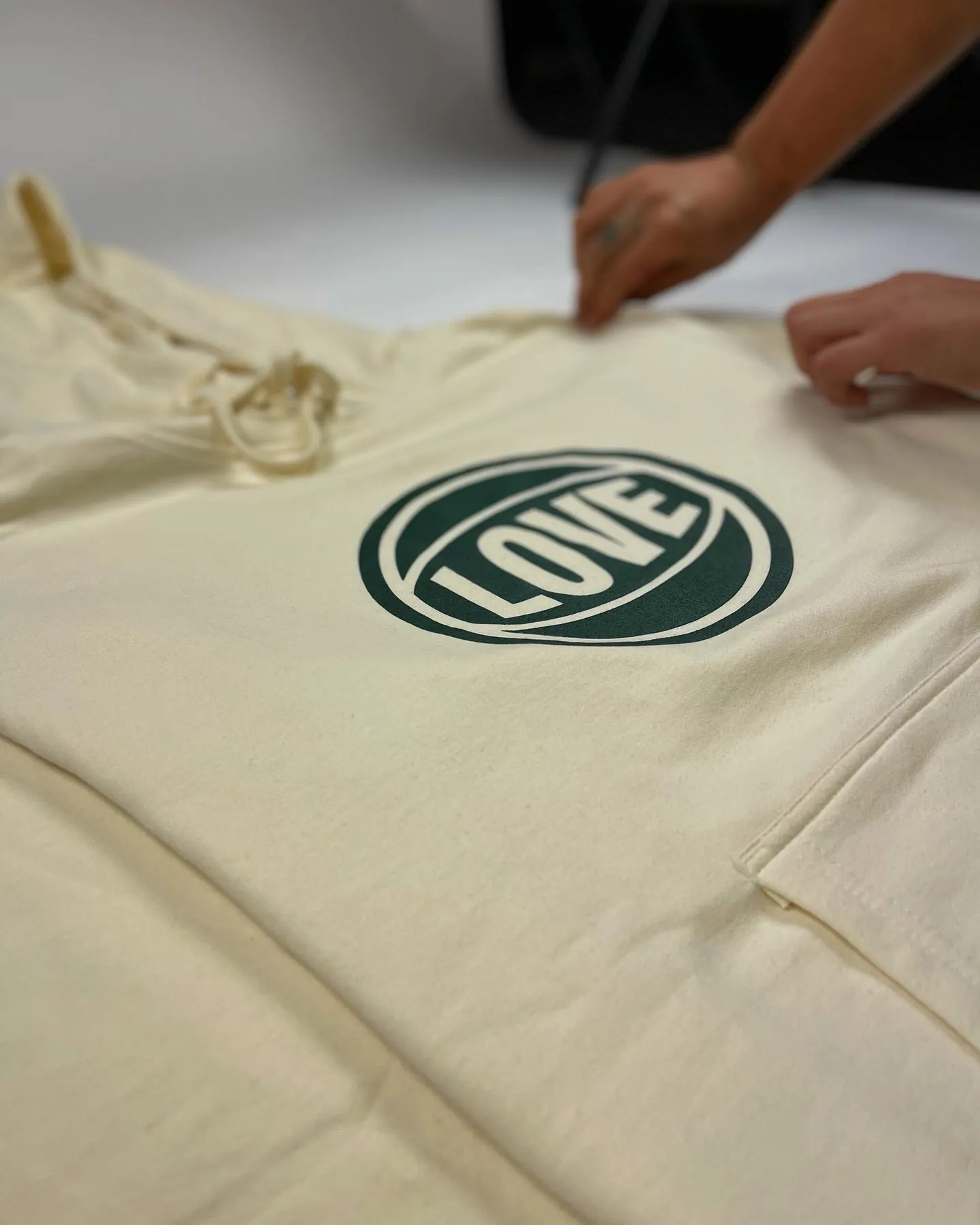 Two hands smoothing a natural cotton hooded sweatshirt laid flat on a work table, screen printed at the chest with a dark green LOVE roundel in a school’s colors.
