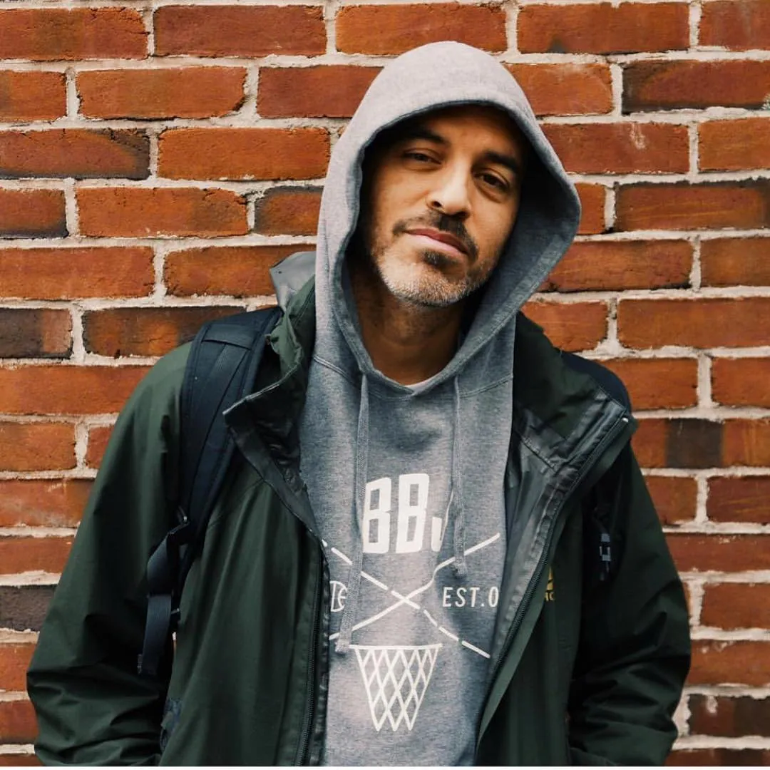 A man in a gray hood pulled up under an open green rain jacket, leaning against a red brick wall and looking at the camera, BBJ printed across the chest of the hoodie over a crossed-lines and net mark